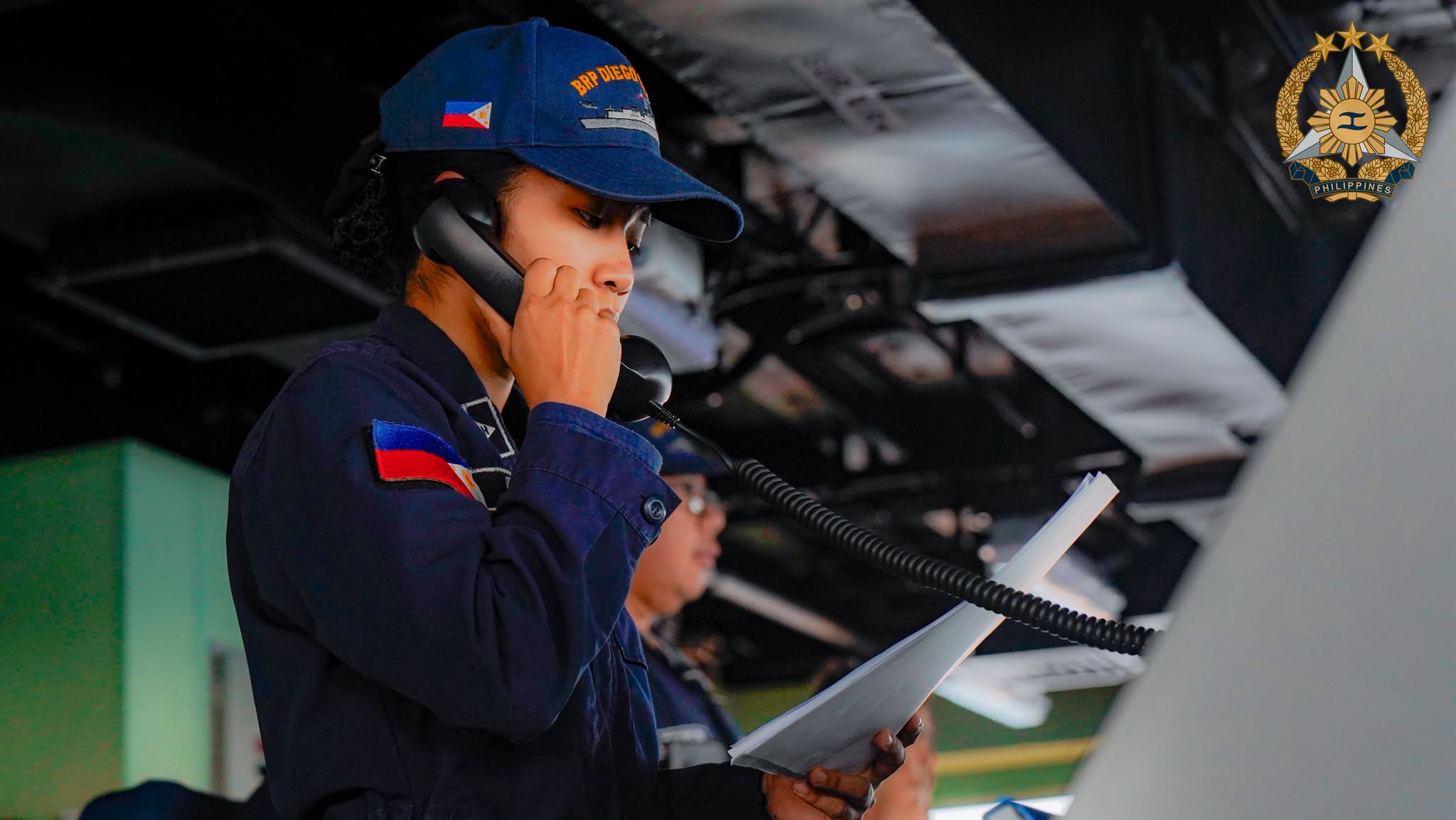 A junior officer on the bridge of the BRP Diego Silang holds a phone handset and a printed script