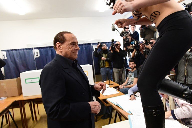 ITALY ELECTIONS. A woman alleged to be a Femen activist jumps on a table in front of Silvio Berlusconi, leader of right-wing party Forza Italia, to protest topless with a body painting reading 'Berlusconi, you have expired' on March 4, 2018, at a polling station in Milan. Photo by Miguel Media/AFP   